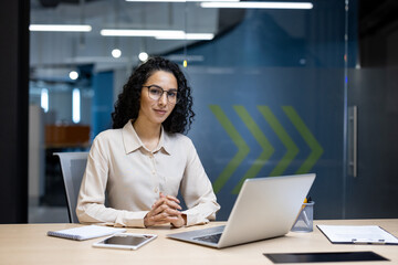 Professional Hispanic businesswoman smiling confidently while seated at her modern workspace, surrounded by digital devices in an office setting.