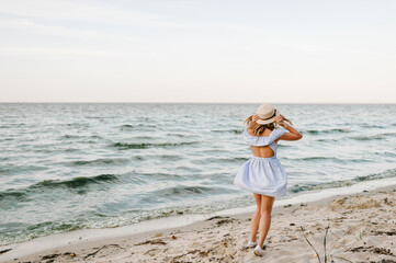 Young attractive blond woman in straw hat joyfully walks near sea. Happy beautiful girl standing on beach ocean and enjoying sunny summer day on vacation. Portrait of stylish female on sand. Back view