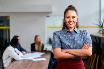 Obraz premium Confident asian businesswoman stands arms crossed in office, team working together in background. Professional leader in smart casual attire, engaging in corporate environment, embraces diversity.