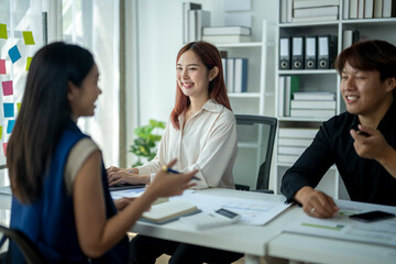 Three people are sitting at a table in a room, smiling and talking to each other. The woman in the center is typing on a laptop, while the other two people are looking at her