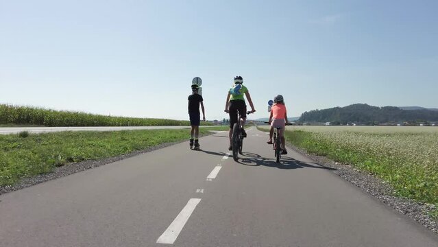 Family sport day in nature, mother with two children enjoying outdoor activity, riding bike and roller skating, rear view. Healthy lifestyle concept.