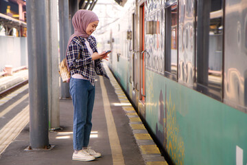 full lenght shoot of happy asian muslim woman holding mobile phone while standing on train station...