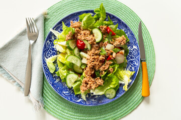 Flat lay view of healthy tuna salad on lettuce, with cucumber and tomato in a vintage plate with blue motif set on round green straw placemat