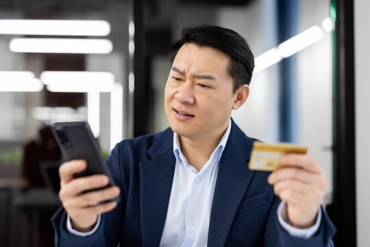 A middle-aged Asian businessman looks confused and concerned while comparing his credit card to his smartphone in an office setting. Concept of financial fraud or banking error.