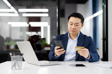 Asian businessman in a blue suit using a smartphone and holding a credit card. He appears focused...