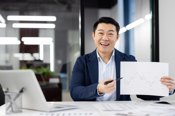 Enthusiastic Asian businessman discussing growth with a financial chart in a modern office setting....