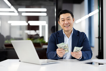 A cheerful Asian businessman counting US dollars at his desk in a well-lit modern office. He exudes...