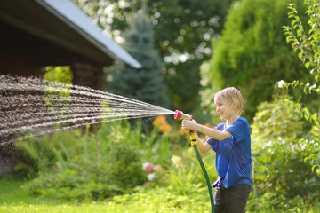 Funny little boy playing with garden hose in sunny backyard. Preschooler child having fun with spray of water. Summer vacation in the village for kids.