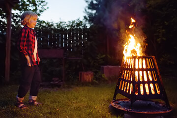 Cute little boy is burning a bonfire on a summer evening in the backyard. Child puts firewood in a...