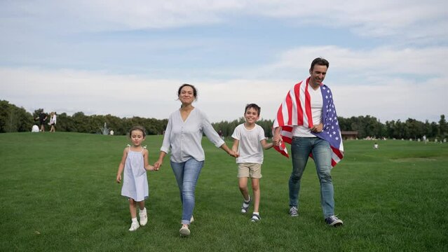 A happy family enjoys leisure time walking in a field with an American flag, surrounded by trees, grass, and a beautiful sky with clouds
