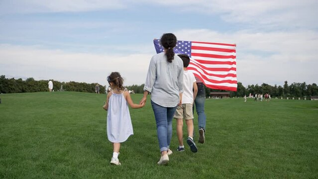 A woman is walking with two children in a grassy meadow, holding an American flag under a cloudy sky. They gesture happily, enjoying leisure time outdoors
