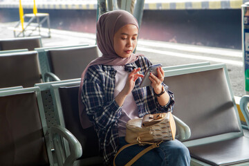 asian muslim woman holding smartphone sitting on bench at train station waiting room. traveling...