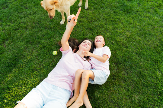Cheerful cute boy kid having fun with his mom, embracing, laughing on the green grass with pet dog in garden on sunset. Summer outdoor activity. Happy active childhood, family time. Selective focus