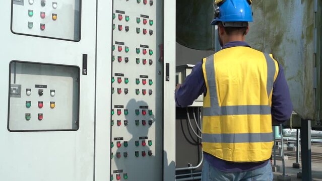 Asian male engineer wearing a reflective jacket, hard hat, walking work electrical cabinet, inspecting electrical systems, building on rooftop of construction building.
