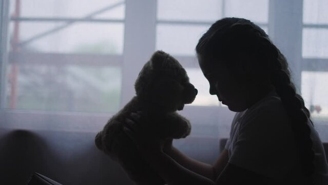 depressed girl child silhouette the window with her favorite toy teddy bear upset. problem of domestic violence in the family concept. depressed girl is sad and lifestyle bored at window with bear