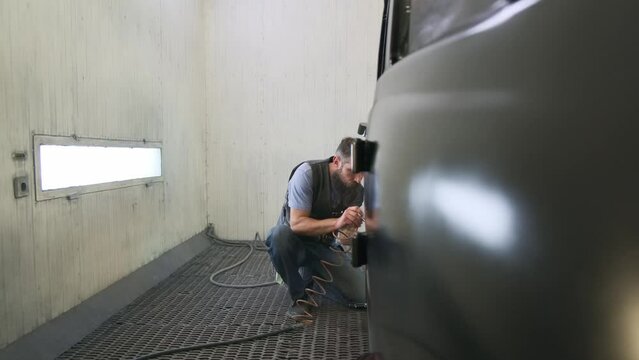 Hot Rod type of custom cars in spray booth. Black painted car in garage corner, master squatted behind airbrushing, long wire. Painting rear surface of car body, camera zooming in, paint technique.