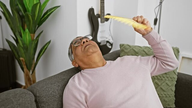Mature man cooling himself with a fan while relaxing on a couch in a living room with a guitar in the background.
