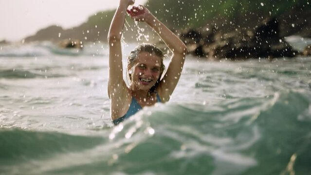 Smiling woman in dress enjoys warm sea, playful splashing water on sunny beach. Romantic carefree solo traveler relishes ocean waves, freedom. Scenic coastal backdrop frames joyful moments. Slowmo