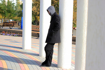 A guy in a coat and hood stands leaning on a column