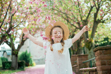 Happy pretty girl 6 years old in white dress in straw hat near blooming pink sakura. The child is...