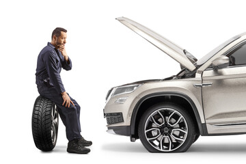 Car mechanic in a uniform sitting on a tire in front of a SUV and thinking