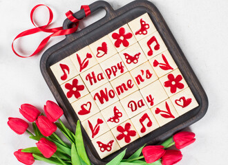 Women's Day. Little square shortbread cookies with marmalade filling of a thematic shape: Happy Women's Day, flower, heart, butterfly, notes. On a wooden plate with a red satin ribbon. With red tulips