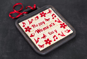 Women's Day. Little square shortbread cookies with marmalade filling of a thematic shape: Happy Women's Day, flower, heart, butterfly, notes. On a wooden plate with a red satin ribbon