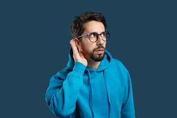 Obraz premium A man with a beard and glasses, dressed in a blue hoodie, stands against a solid color background. He appears focused as he cups his hand behind his ear, trying to hear something more clearly.