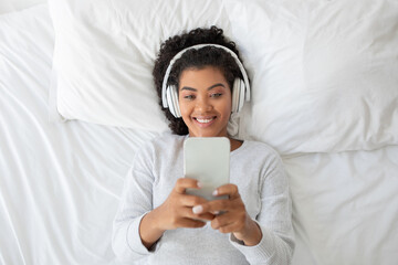 Hispanic woman lying down on a bed, actively engaged in listening to music through her smartphone....