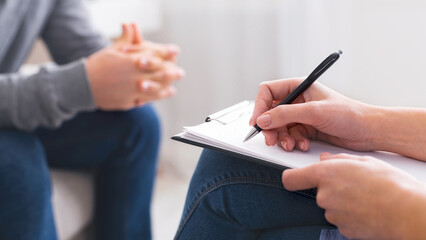 A close-up view showcasing a therapists hands holding a pen and writing on a notepad, with a patients hands in the background gesturing mid-conversation, cropped