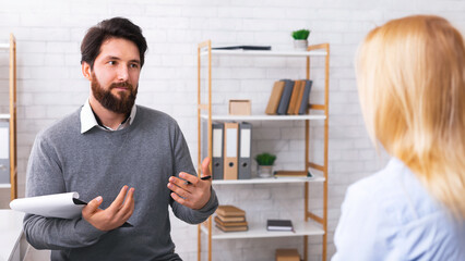 Bearded millennial man consulting blonde woman client in cozy office interior, holding clipboard...