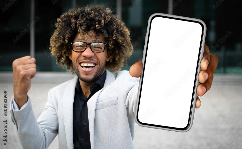 Poster African American man dressed in a suit standing while holding up a cell phone, looking at the screen intently. He appears focused and engaged in whatever is displayed on the device.