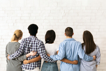 A group of multiethnic teenagers stands close together with their backs to the viewer, arms around each other in a display of friendship and unity, back view
