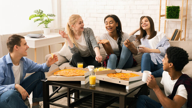 A diverse group of multiethnic friends are gathered around a wooden table. They are enjoying slices of hot pizza, engaged in casual conversation and laughter. - Powered by Adobe