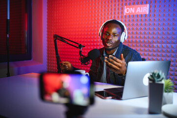 african american vlogger using smartphone to film podcast in studio. blogger with mobile phone, microphone and headphones filming video