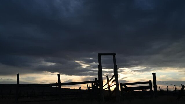 Time lapse of dark late day clouds with a layer of bright sunset sky behind an old horse or cattle corral in the foreground. The foreground is in silhouette.
