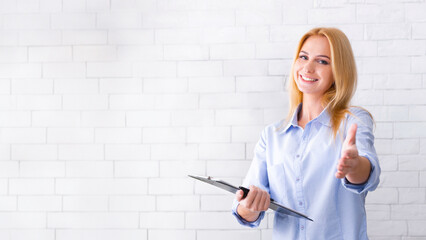 A smiling professional businesswoman stands against a white brick wall holding a clipboard in one...
