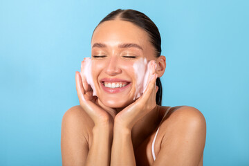 Cheerful laughing woman applying cleansing foam for washing face, blue background. Skincare and beauty routine concept