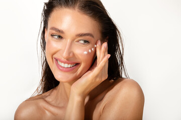 Beautiful lady with wet hair applying eye cream and touching cheeks, looking at free space and...