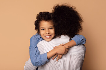 A young African American boy with curly hair smiles brightly as he embraces a woman, presumably his...