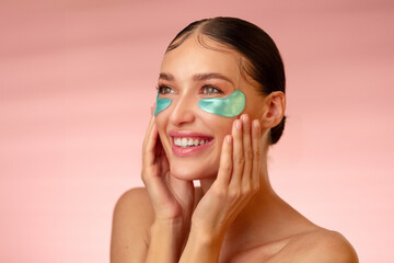 Femininity and skin care. Young European woman applying eye patch and smiling while touching cheeks, standing against gradient peach background