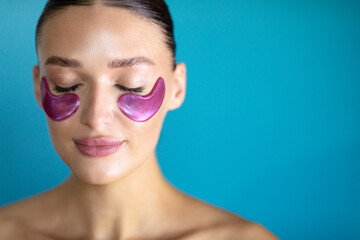 Attractive half-naked lady with closed eyes applying purple eye patches under eyes, posing isolated on blue background, free space
