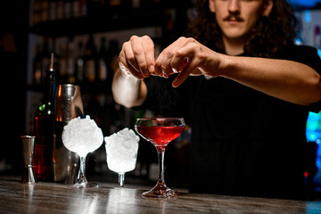 Bartender's hands squeeze a piece of zest, holding it directly above a glass with a brown cocktail