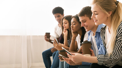 A group of multiethnic teenagers is seated close to one another, each engrossed in their own...