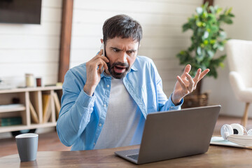 A man appears visibly upset as he argues over the phone, gesturing with his hand. He is seated at a wooden table where his laptop is open, suggesting he is dealing with a work-related issue