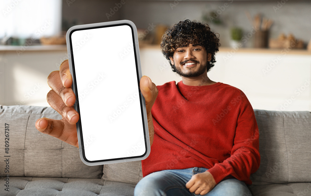 Poster eastern man is seated on a couch, holding a smart phone in his hand. he appears focused on the devic