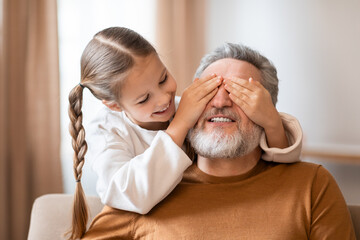 A young girl with a ponytail is covering her grandfathers eyes from behind as they sit together,...