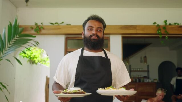 Indian waiter in apron carries food to cheerful diverse women in cafe on Bali. Friendly waiter smiling, talking with female customers in the tropical restaurant. Charming waiter serving clients.