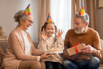 A young girl wearing a party hat is sitting between her smiling grandparents, who are also adorned...