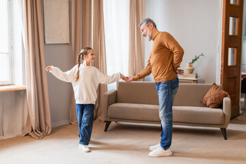 A cheerful grandfather is engaging in a playful dance with his young granddaughter in the warm and...